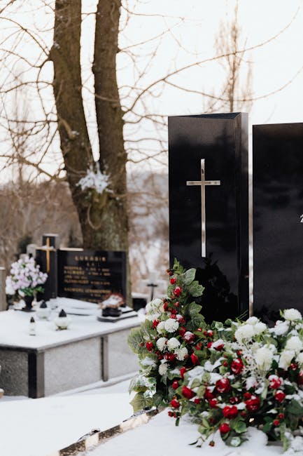 A floral arrangement on a funeral grave featuring a lush bouquet of red and white chrysanthemums, surrounded by green foliage, positioned at the base of a black granite headstone with a silver cross. 