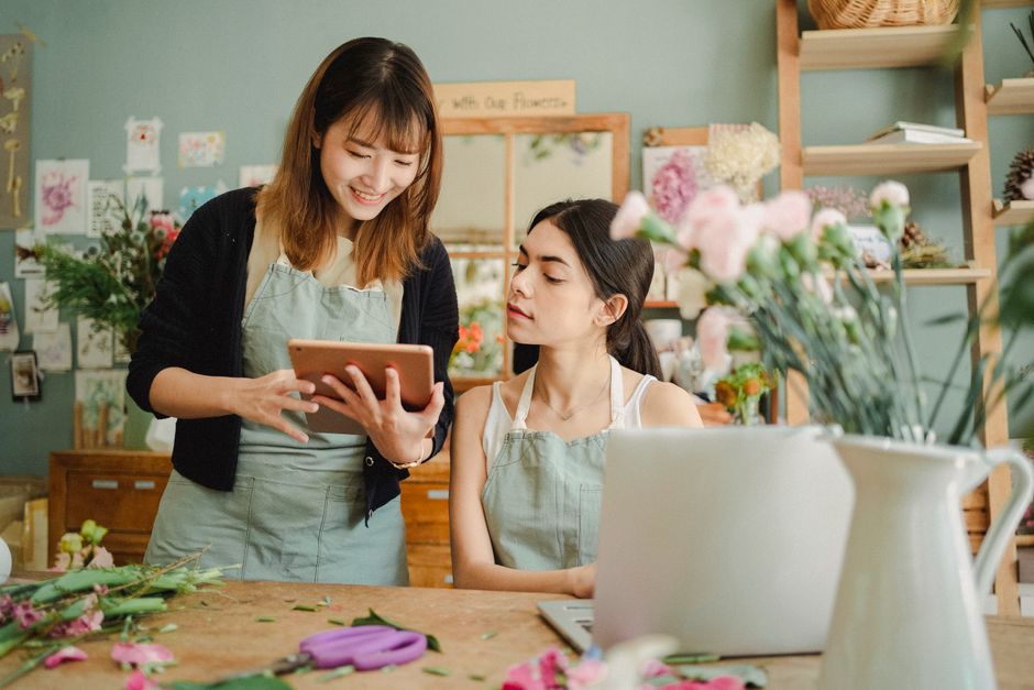 Two women working in a floral workshop, one standing and holding a tablet and the other seated. The standing woman has shoulder-length brown hair, is smiling, and wearing a black cardigan over a green