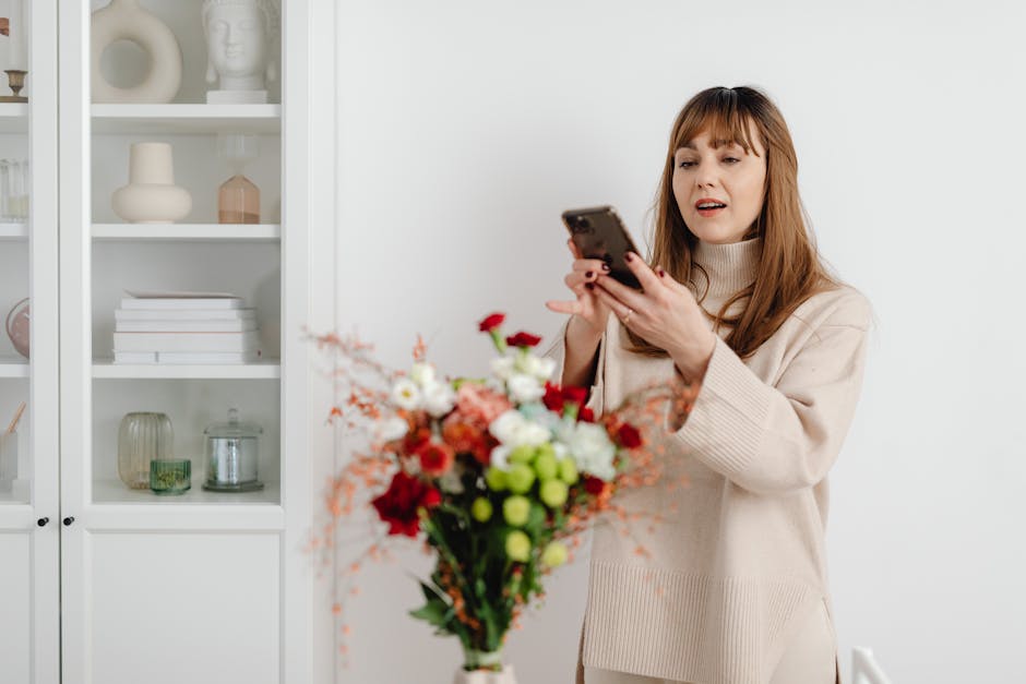 A woman with long, light brown hair and bangs, wearing a beige oversized sweater, stands indoors holding a smartphone in her right hand and a colorful floral bouquet in her left hand. The bouquet feat