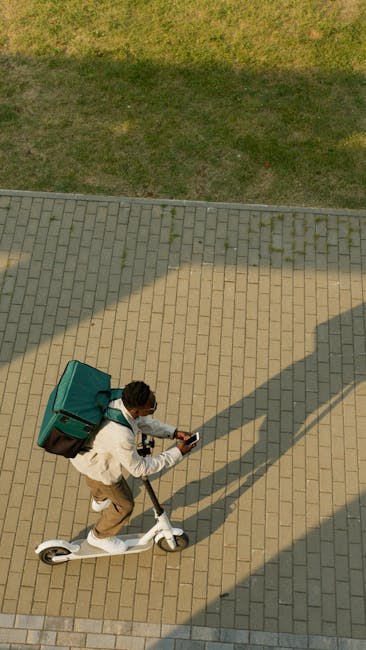 An overhead view of a person riding a white electric scooter on a paved pathway, carrying a large teal delivery box on their back. The pathway is composed of rectangular beige and gray bricks, with th