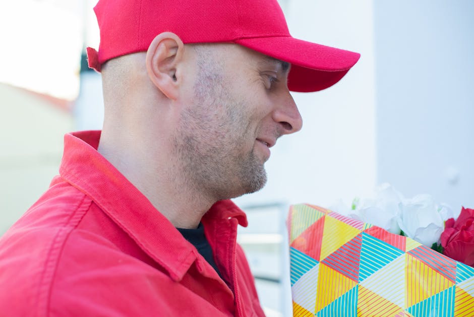 A man wearing a red baseball cap and a matching red jacket is holding a colorful, geometric-patterned gift bag filled with white and red flowers. The bouquet features fresh petals and green stems, arr