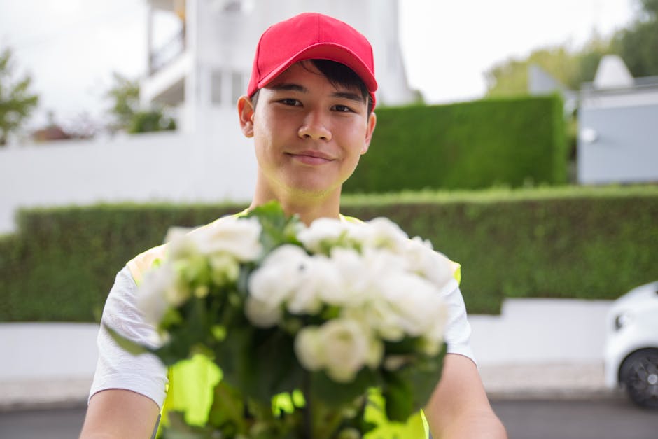 A delivery person wearing a red cap and a high-visibility vest is smiling and extending a bouquet of fresh white flowers towards the camera outdoors. The bouquet features white roses and other small w