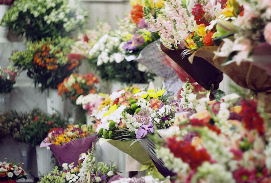 A vibrant display of floral arrangements featuring a variety of fresh flowers including pink, white, yellow, orange, and purple blooms, wrapped in colorful paper and elegant ribbons. The flowers are a