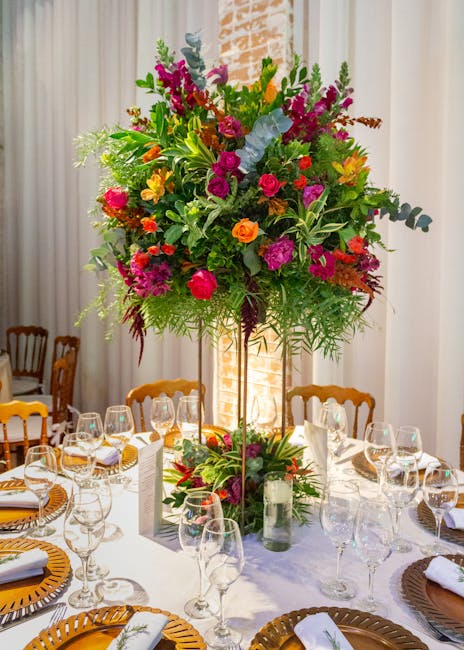 A large, vibrant floral centerpiece featuring an assortment of fresh flowers in bright pinks, purple, orange, yellow, and green foliage, arranged in a tall, slender glass vase. The bouquet includes ro