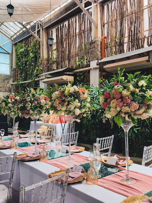A long banquet table decorated for a wedding setting with elegant floral arrangements. The arrangements feature large, lush bouquets of soft pink, white, and cream roses, peonies, and hydrangeas, acce